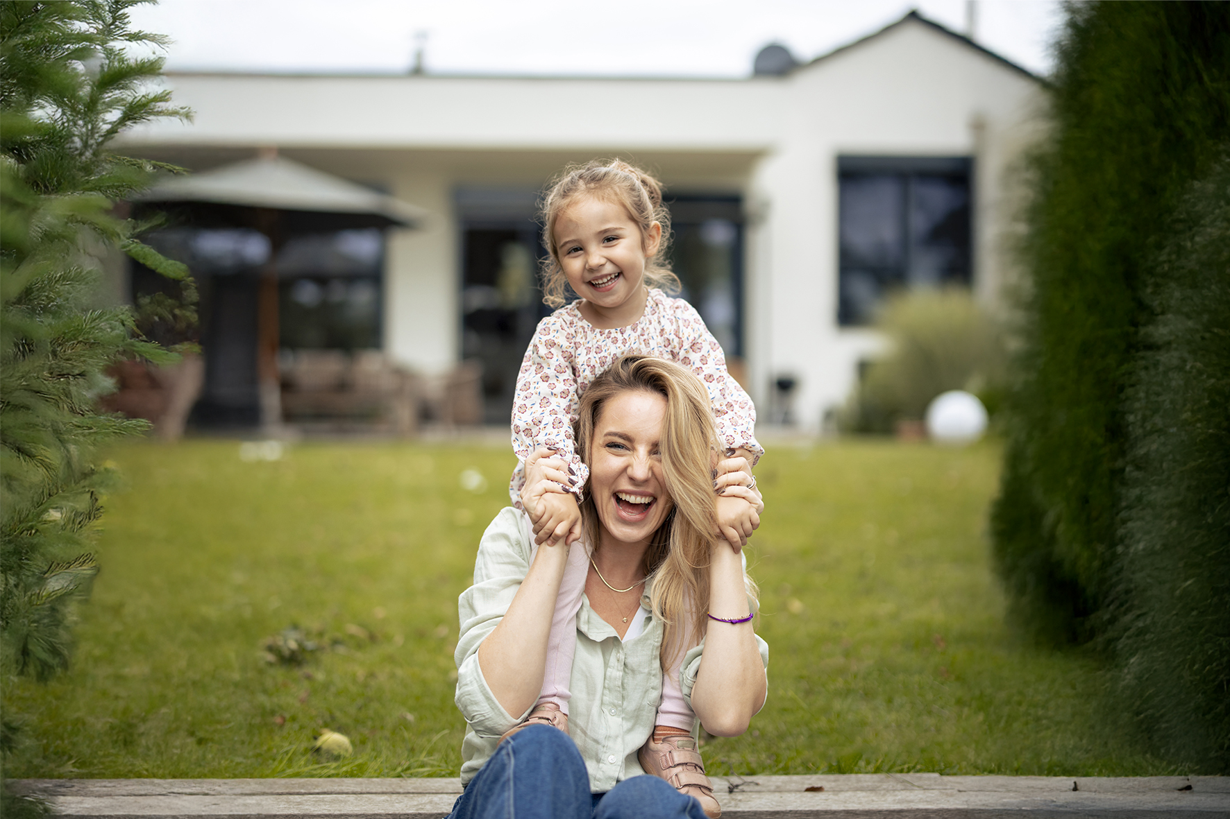 Mutter und Tochter im Garten vor Haus. UniVersa Wohngebäudeversicherung schützt das Eigenheim
