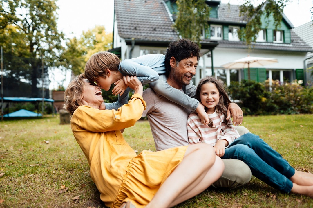Familie tobt im Garten, Eigenheim im Hintergrund. Versicherungsempfehlungen, um sich und die Familie richtig zu versichern