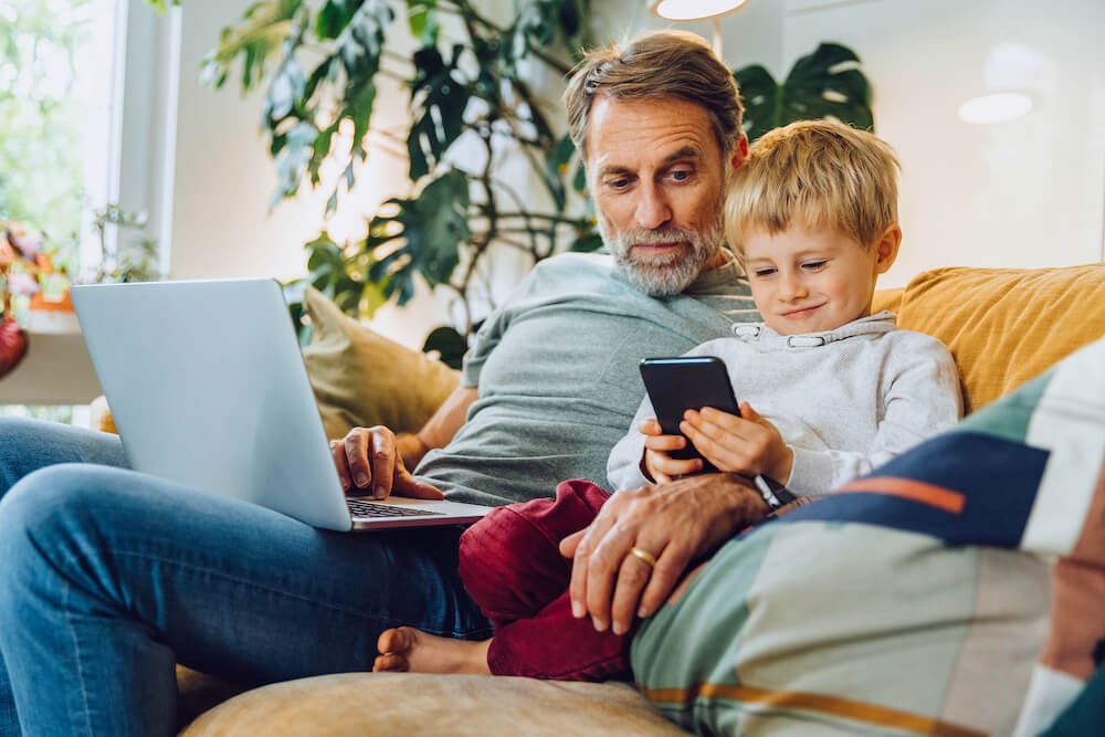 Mann mit Laptop sitzt mit Jungen und seinem Handy auf Sofa. Symbol für den universa Tarifrechner für Jung und Alt
