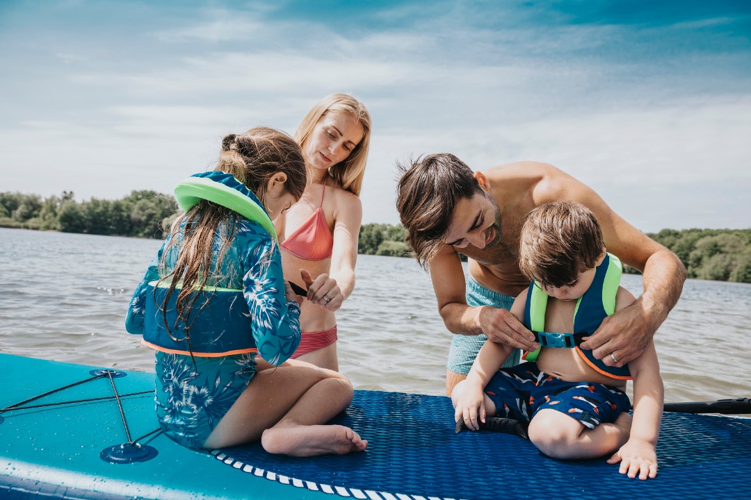 Junge Familie am Badesee, Eltern ziehen Kindern auf SUP Schwimmwesten an. Universa Kindervorsorgekonzept für sorglose Zukunft